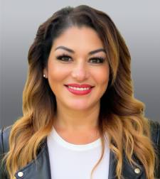 Portrait of a smiling woman with long, wavy brown-to-blonde balayage hair, wearing a white top and dark jacket, against a gray gradient background.