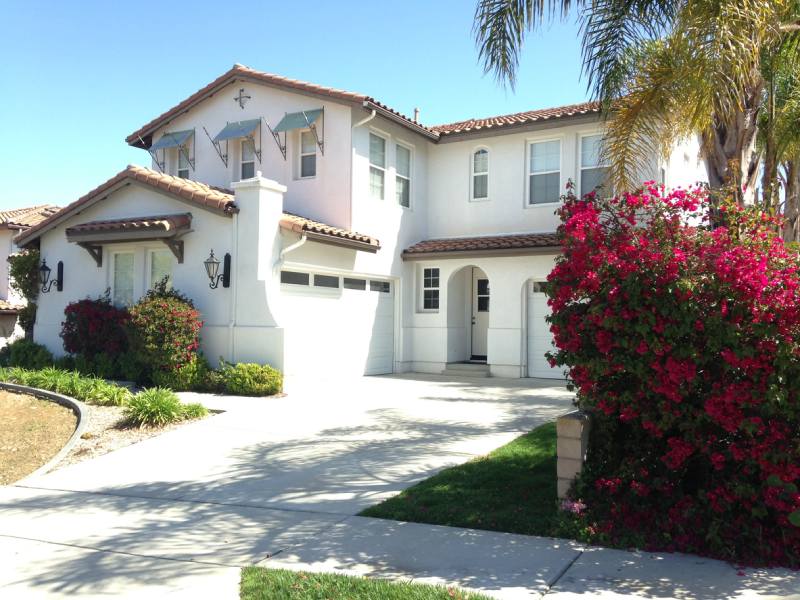 White two-story stucco house with a red-tile roof, palm trees, and a flowering pink bush beside the driveway.